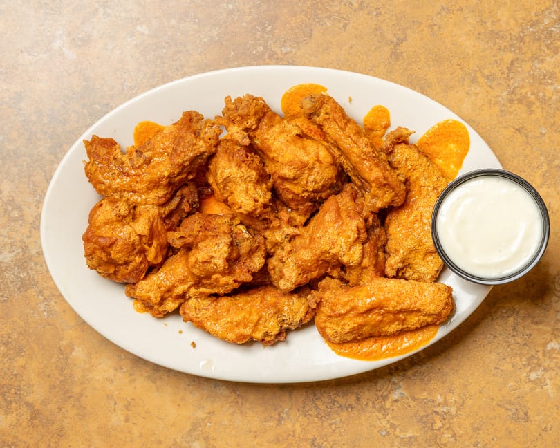 A white oval plate filled with golden-brown chicken wings, drizzled with orange sauce, and a side of ranch.