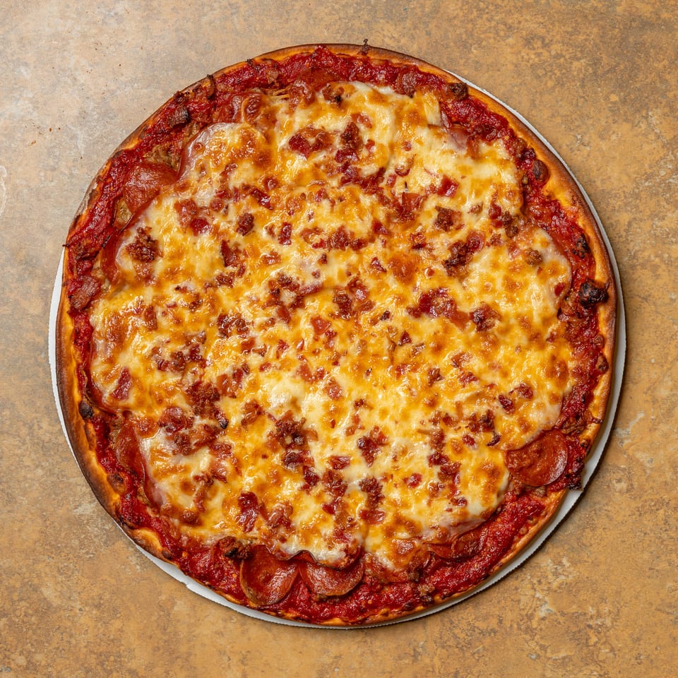 A whole pepperoni and bacon pizza with melted cheese and a thick crust on a brown surface.