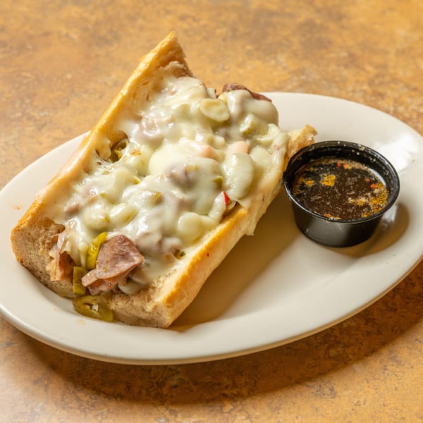 A cheesy Italian beef sandwich with giardiniera and a side of au jus on a white plate.