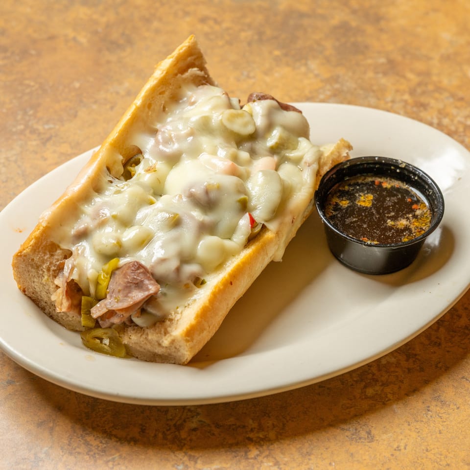 A cheesy Italian beef sandwich with giardiniera and a side of au jus on a white plate.