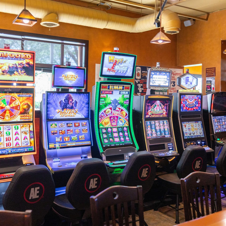 A row of brightly lit slot machines with various games and chairs in a casino or bar.