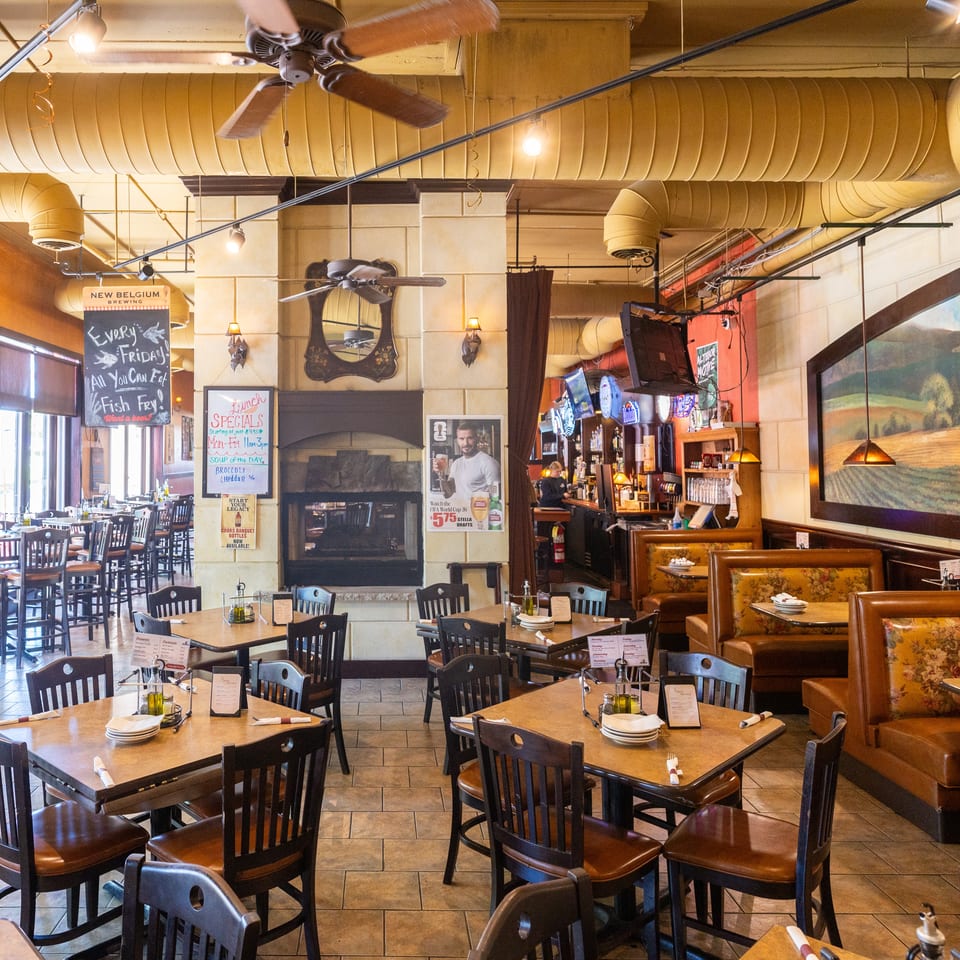 An empty restaurant dining room with tables, chairs, booths, a bar, and a large landscape painting.