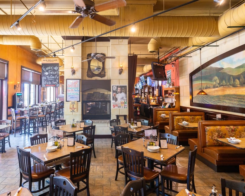 An empty restaurant dining room with tables, chairs, booths, a bar, and a large landscape painting.