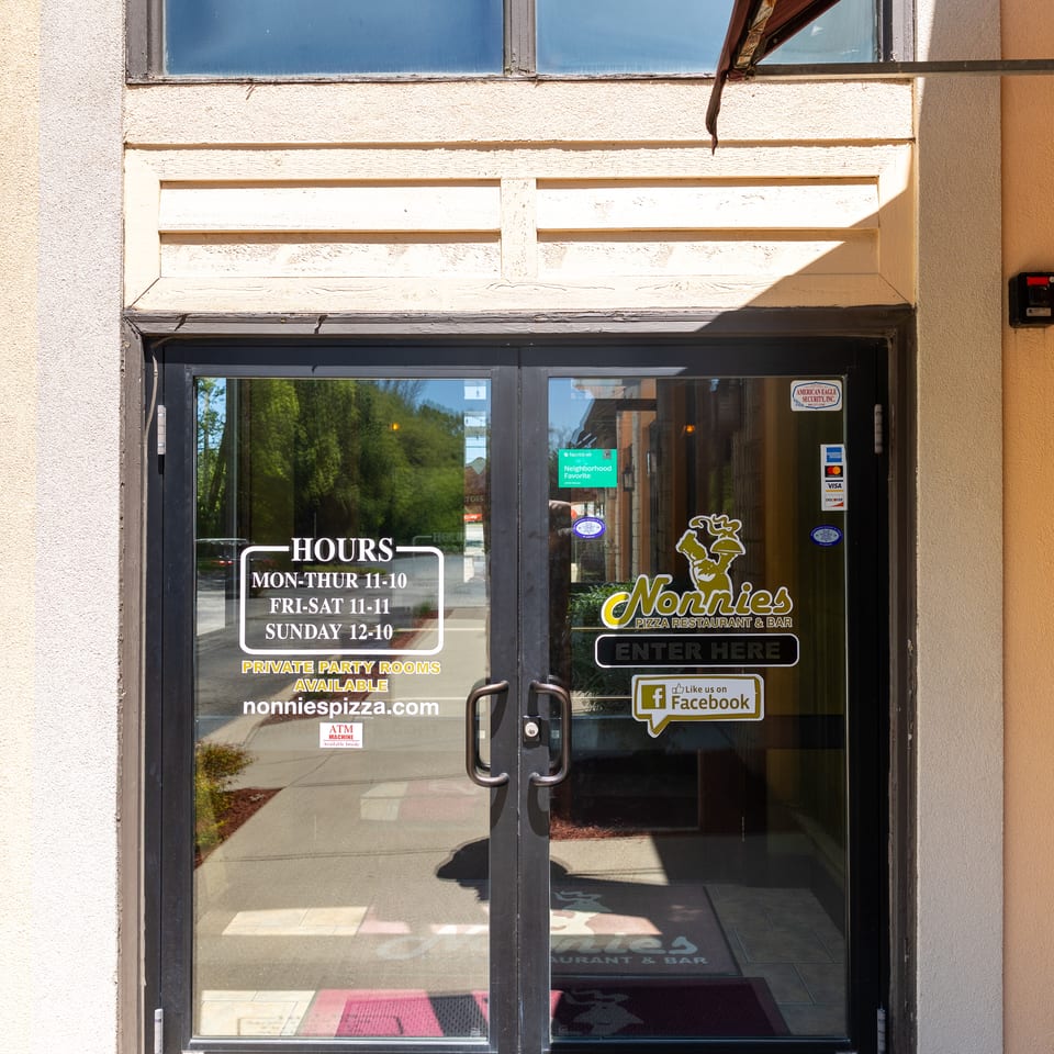 The glass entrance doors of Nonnies Pizza Restaurant & Bar, displaying hours, website, and social media.