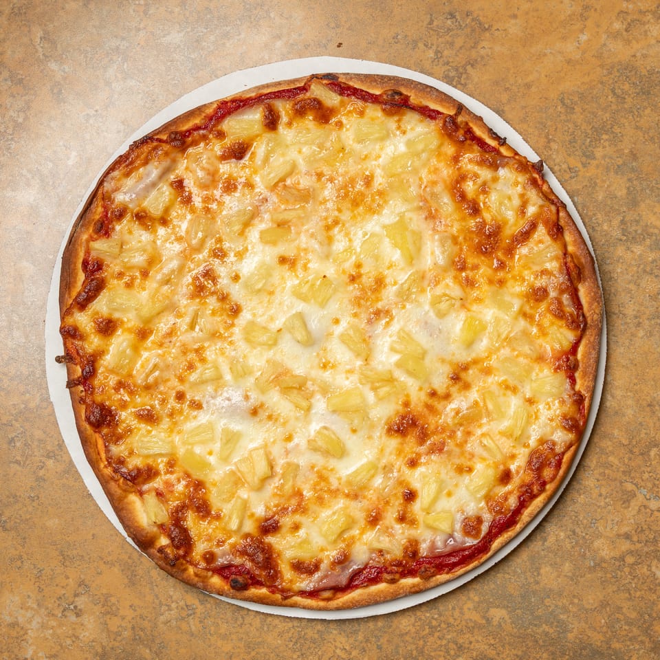 A whole pineapple pizza with melted cheese and tomato sauce on a brown textured surface.