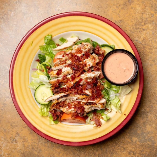 Grilled chicken salad with bacon, lettuce, tomatoes, cucumbers, and a side of dressing.