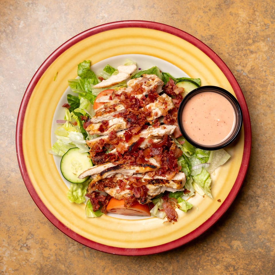 Grilled chicken salad with bacon, lettuce, tomatoes, cucumbers, and a side of dressing.